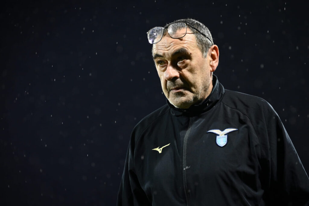 Maurizio Sarri in primo piano con la giacca della Lazio, preoccupato per il blocco del mercato
